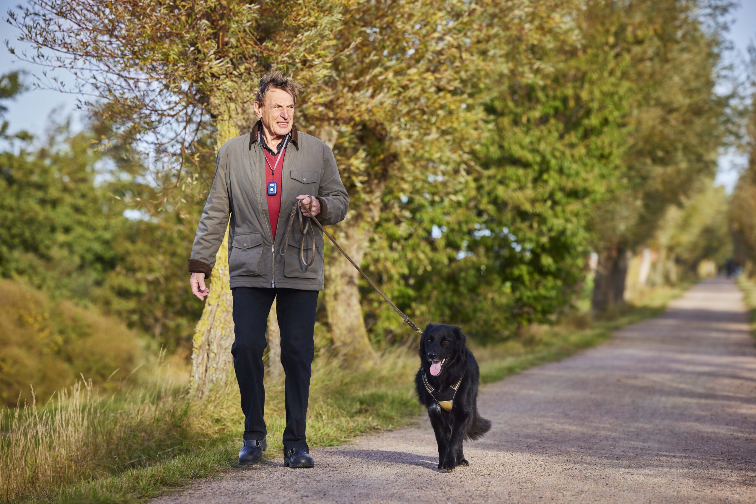 Äldre man med ett mobilt trygghetslarm på promenad med en svart hund i naturen