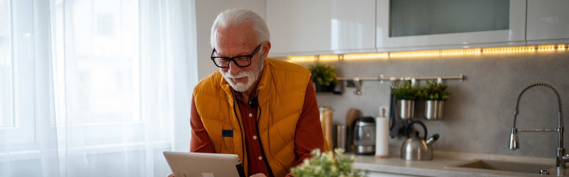 Äldre man med gul väst står i ett kök och har ett digitalt besök med en vårdgivare