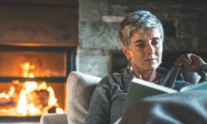 Femme âgée assise sur un canapé lisant un livre avec une cheminée en arrière-plan
