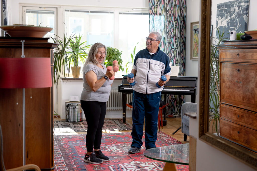 an elderly man and woman holding dumbbells in a living room while being remote monitored by camera visit