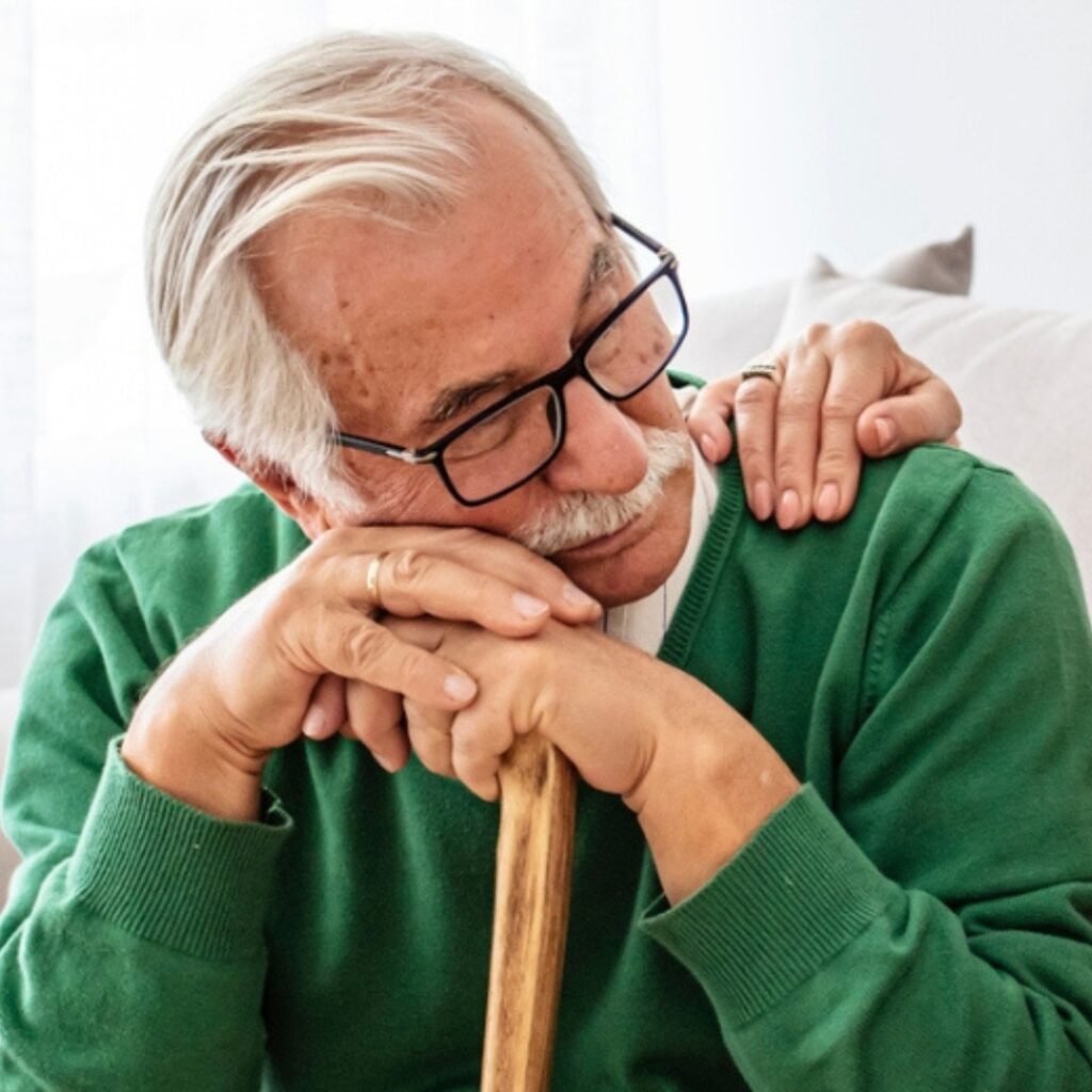 elderly man sitting down, leaning on a cane and wearing a green sweater