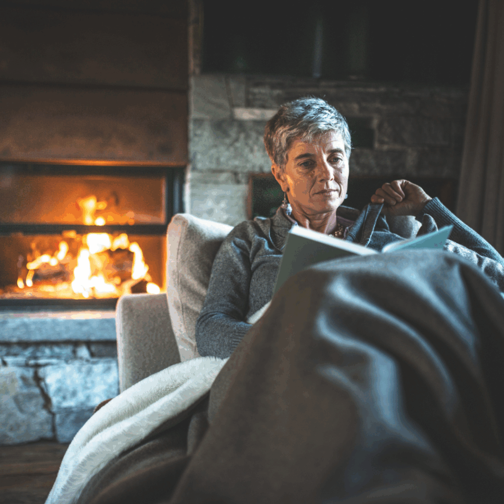 Mujer mayor leyendo un libro con una chimenea al fondo.