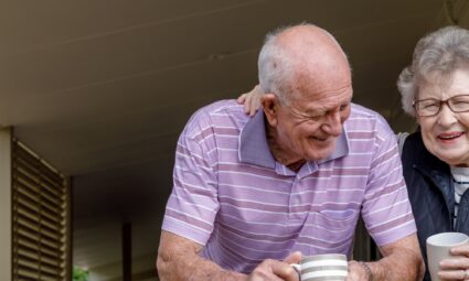 old couple standing on a porch drinking coffee and laughing
