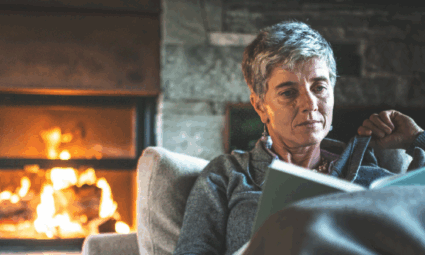 Older woman sitting in a sofa, feeling cozy and reading a book with an open fireplace in the background