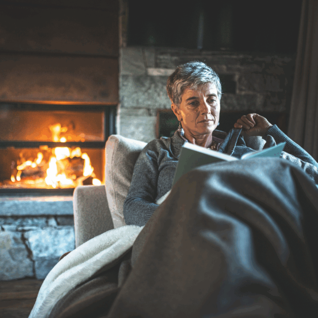 Older woman feeling cozy and reading a book with an open fireplace in the background