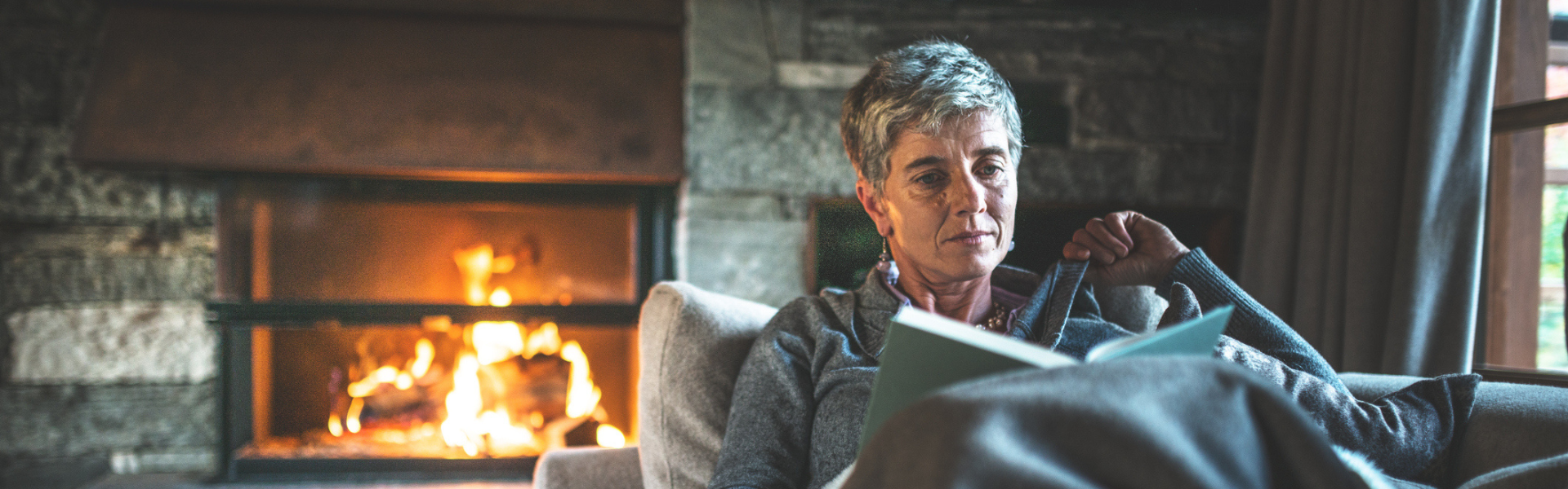Older woman sitting in a sofa, feeling cozy and reading a book with an open fireplace in the background