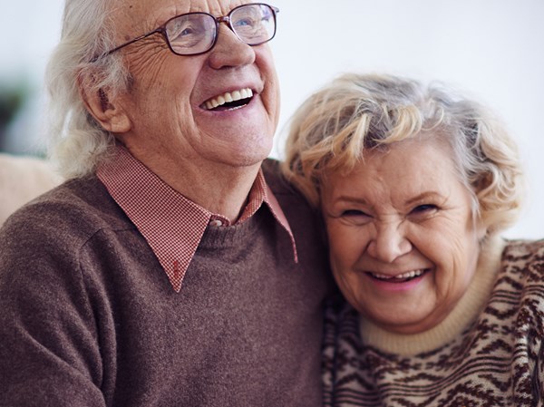 Zwei ältere Menschen sitzen lachend auf einer Couch und halten sich an den Händen