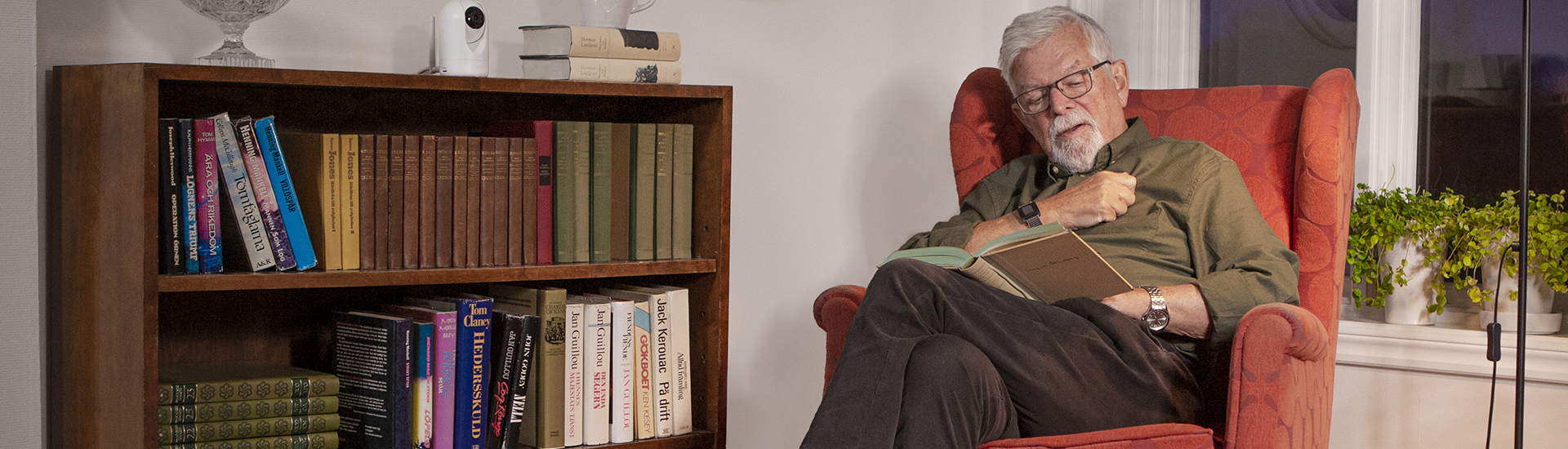 Eldre mann i lenestol som leser en bok, og Doro Visit-kamera på bokhyllen 