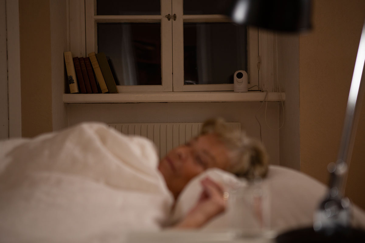 Äldre kvinna sover i en säng med Visit i bakgrunden.