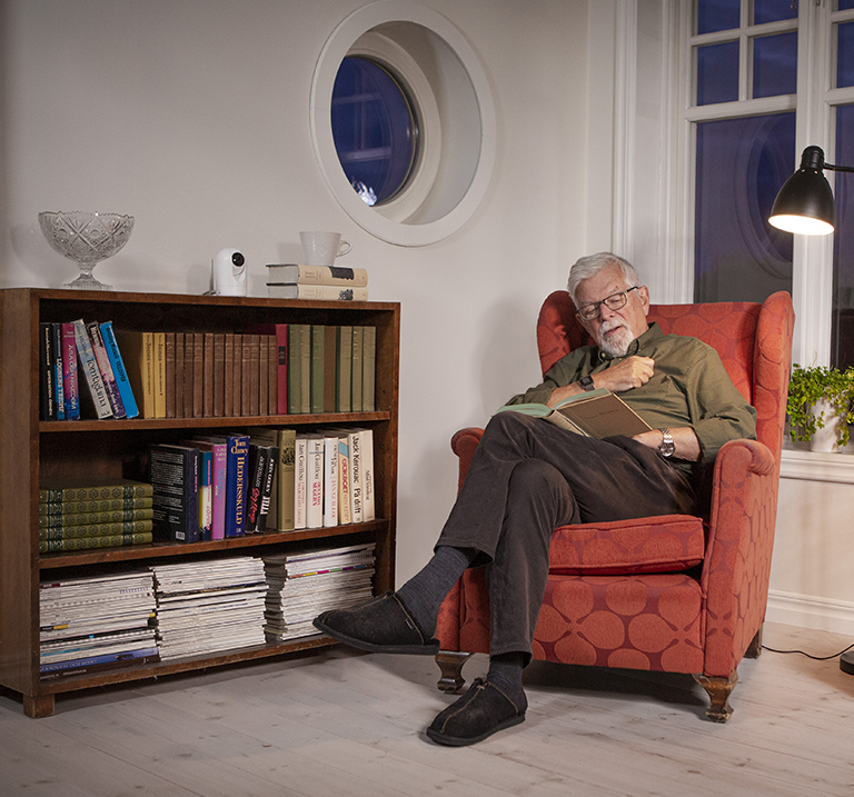 Älterer Mann im Sessel, ein Buch lesend, die Doro Visit-Kamera im Bücherregal 