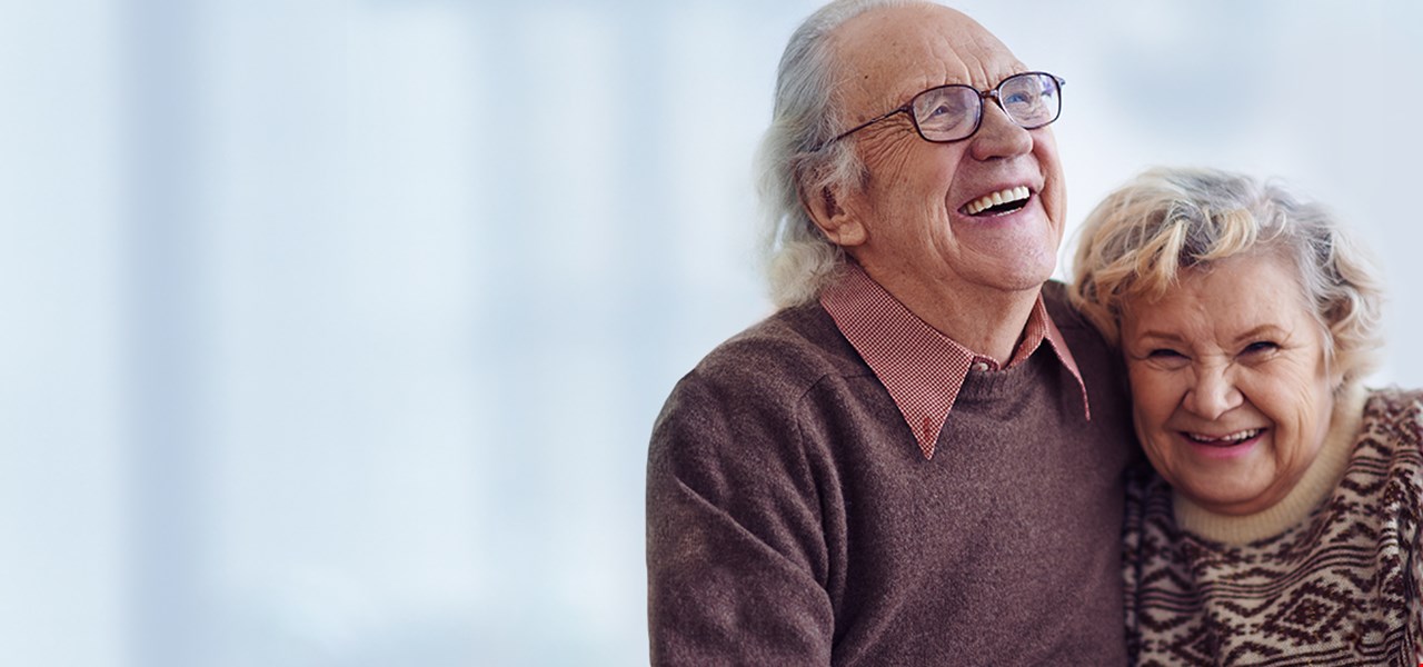 To eldre som sitter på en sofa og ler og holder hverandre i hånden