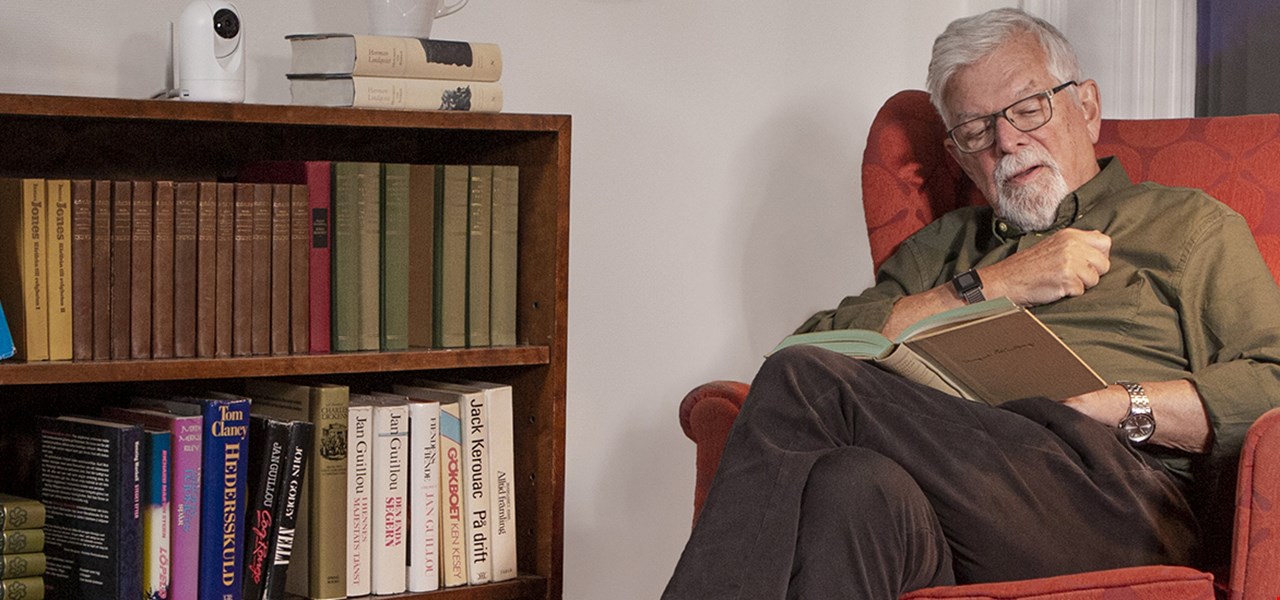 Äldre man i fåtölj som läser och en Doro Visit-kamera ovanpå en bokhylla